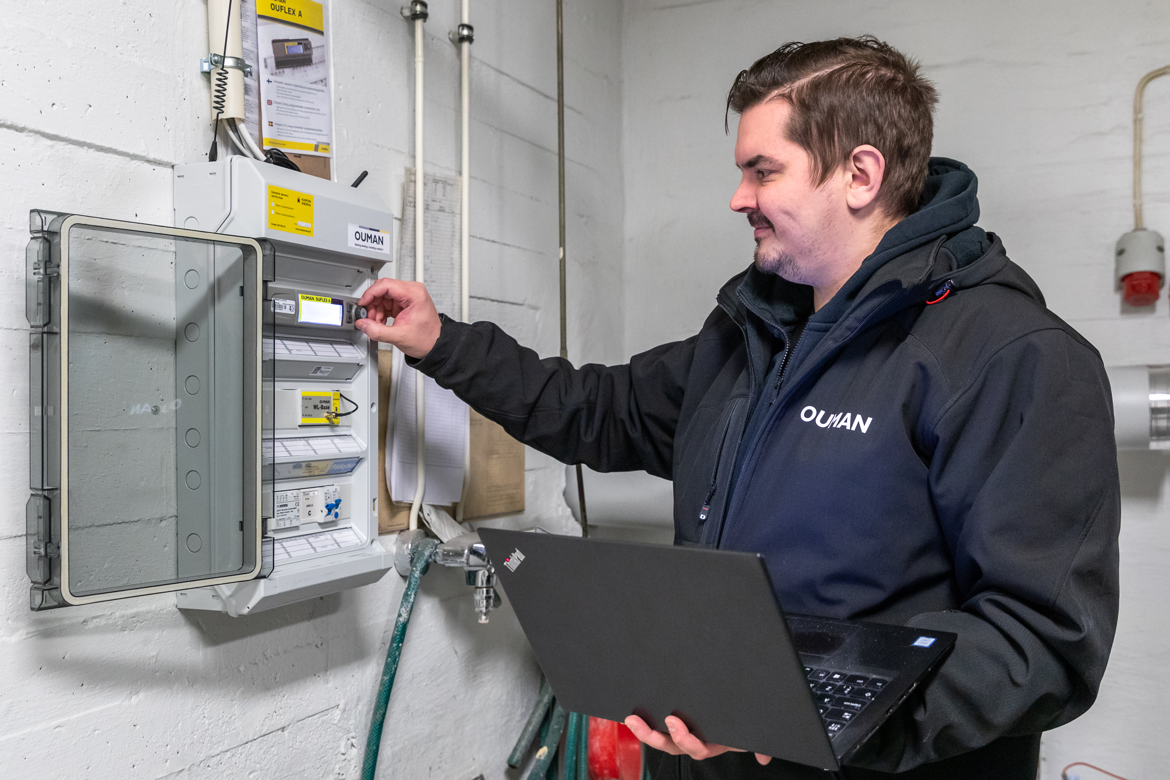 Technician adjusting an Ouman controller in an electrical cabinet while holding a laptop.